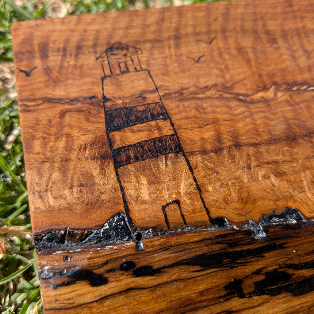 Tabla de servir El faro, hecha a mano por Siete Maderas en madera de roble, usando el nudo como parte del diseño y con detalles en resina simulando el mar y pirograbado dibujando un faro y unas gaviotas. Detalle el faro.