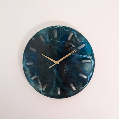 Reloj de pared redondo de resina Skuld, azul oscuro con vetas blancas que evocan el mar profundo. Agujas doradas, diseño abstracto. Una decoración única para cualquier espacio.